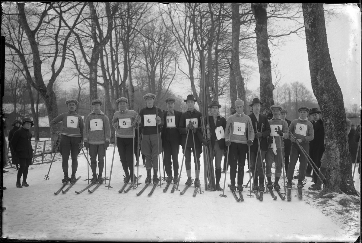 Deltagare i Alingsåsmästerskapen på skidor på sträckan 1 mil. 11 män är uppställda på rad, med nummerlappar på bröstet och skidor på fötterna. Åskådare syns på båda sidor om dem.