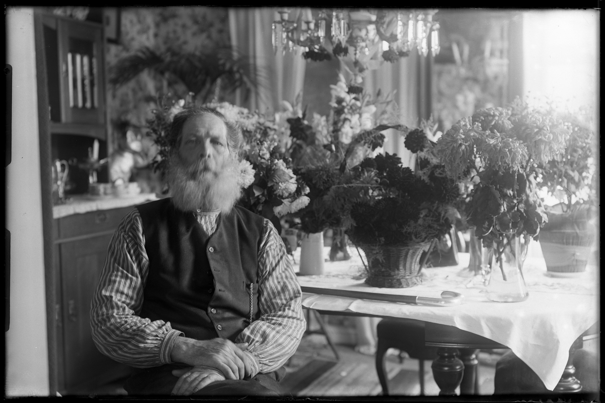 En man i randig skjorta och väst sitter vid ett bord fullt av blommor. I fotografens egna anteckningar står det "Lycke 75 år"