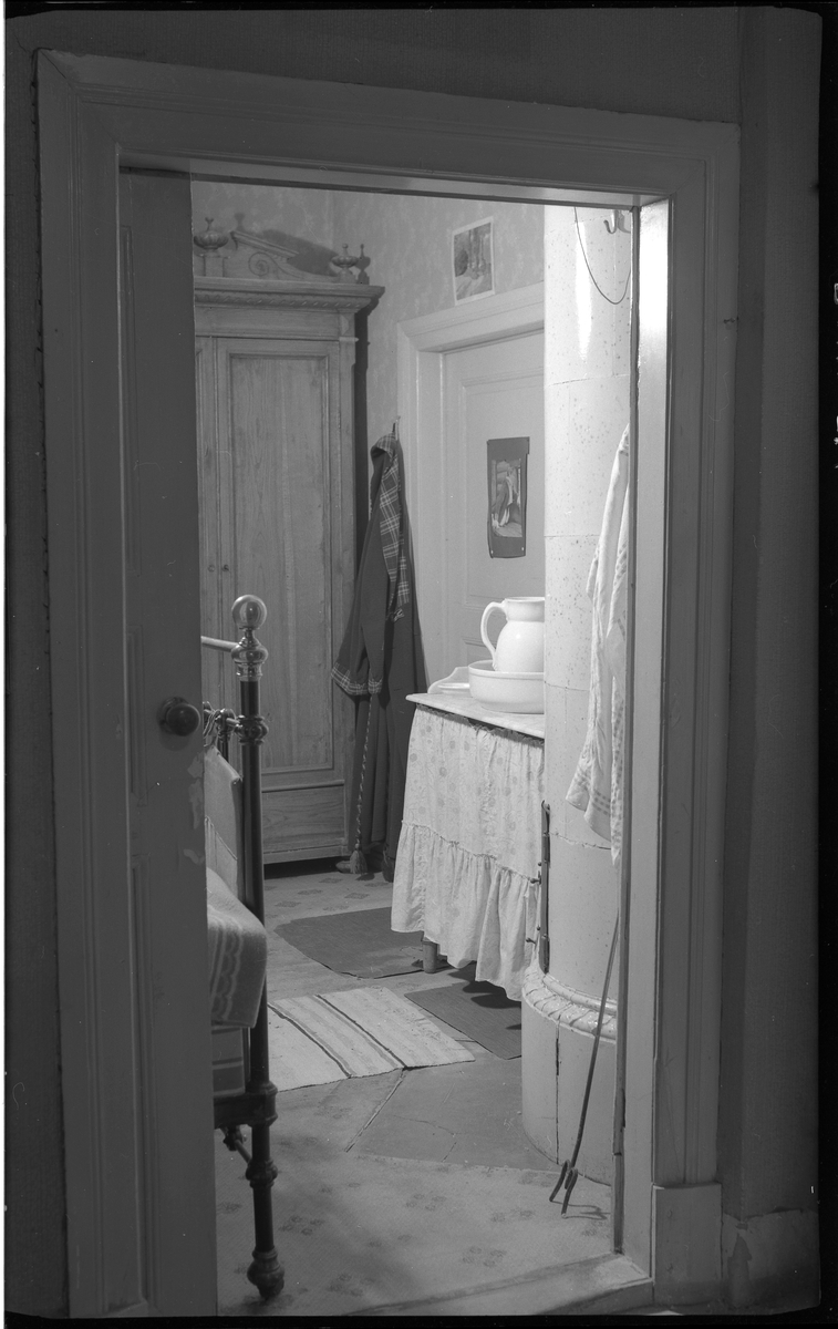 Interiör från en äldre bostadshus, villa Charlottenberg (Hedvigsberg) vid Göteborgsvägen. Huset byggt på 1800-talet.

Genom en öppen dörr ser man in i ett sovrum. Till vänster innanför dörrkarmen syns gaveln av en järnsäng.

I rummet står ett högt skåp av trä, krönt med dekorerad front.
Ett toalettbord med kappa och med ett tvättfat med tillhörande kanna.
En morgonrock hänger på en krok mellan högskåpet och en stängd dörr.

På golvet ligger trasmattor.

På andra sidan dörröppningen syns delar av en rund, vit kakelugn.
En eldgaffel står lutad bredvid.

Elin Bothén.

Foto 13 september 1966.