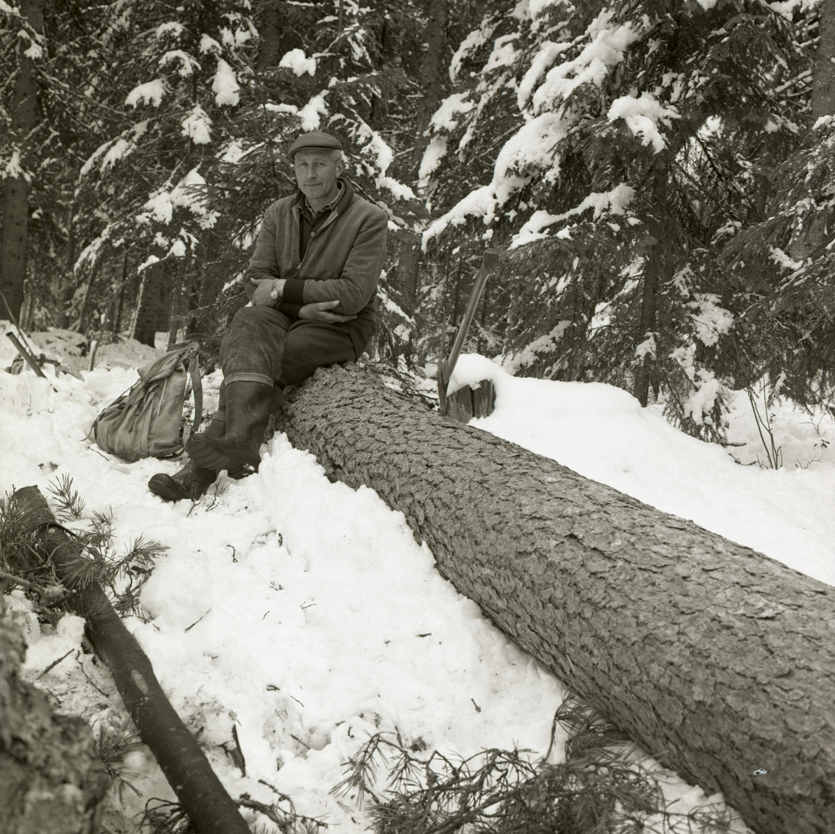 En man sitter på en liggande trädstam i skogen och intill honom ligger en ryggsäck i snön.