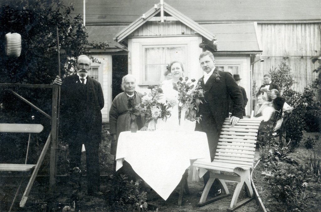 Alfred Träff och hans fästmö vid deras förlovningsfest. Paret står tillsammans med ett äldre par runt ett bord med buketter i en trädgård. Bakom dem syns ett hus och flera personer.