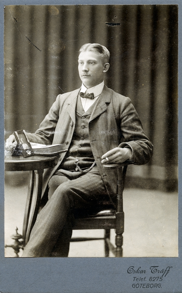 Ateljéporträtt av Oskar Träff. oskar sitter i en stol vid ett litet bord på vilket det ligger ett fotoalbum och står ett inramat fotografi med ett kvinnoporträtt. Oskar har högra handen vilandes i albumet och i vänstra handen håller han en cigarett.