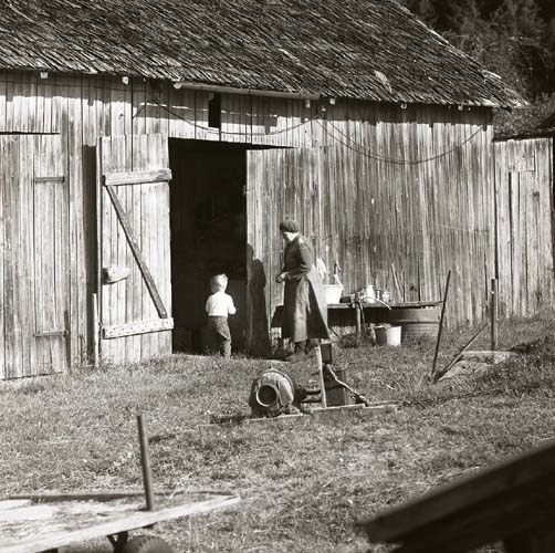 En kvinna och ett barn står vid en ladugårdsdörr. Bakom dem ligger en elmotor på marken, 1969.