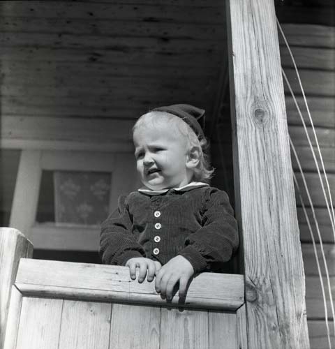 Ett barn iförd mössa och jacka står på en förstukvist och håller i ett räcke, 1950.