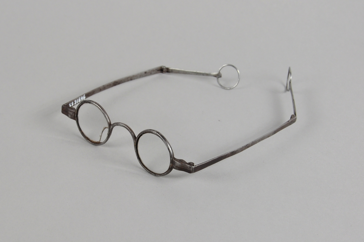 Runde briller med metallinnfatning. Stengerne kan brettes ut, og har en ring ytterst. Det ene glasset er knust.