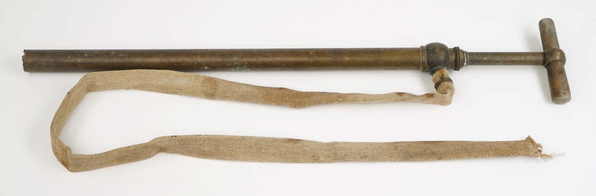 Messingrør med håndpumpe med håndtak av dreid tre. Til røret er det feste en slange av tekstil.