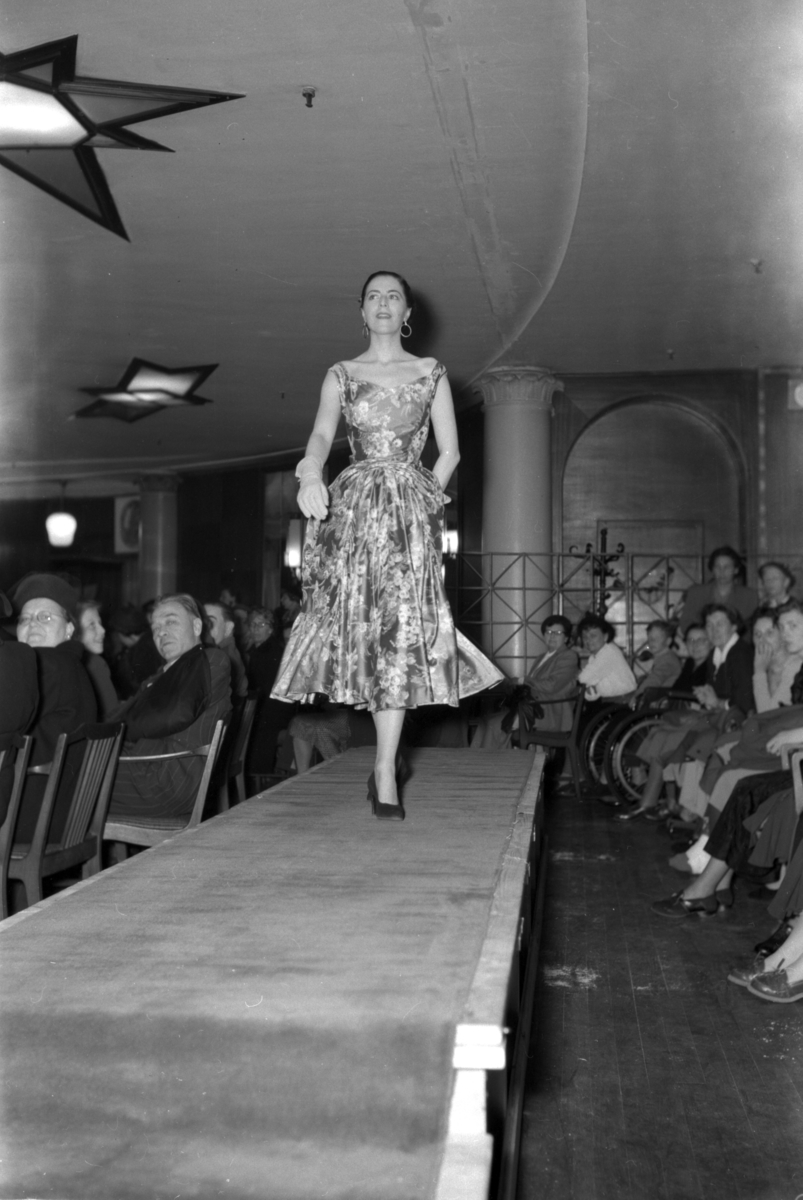 En kvinne på podiet iført mønstrete kjole. Fra en motevisning på Steen & Strøm fra 7. mai 1954..