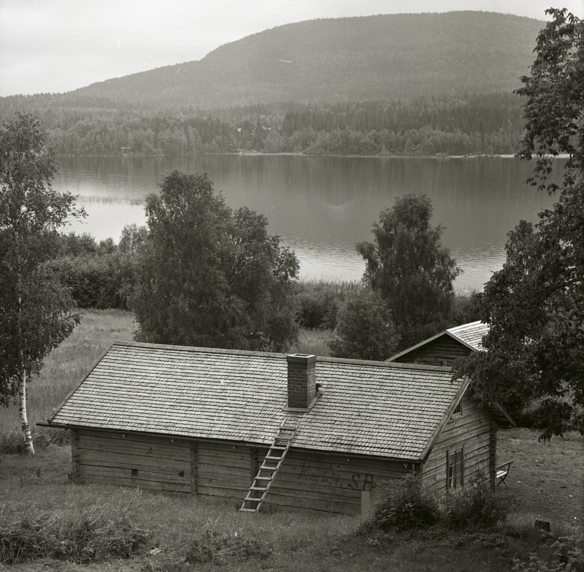 En utsikt över en fäbodstuga med en sjö och ett berg i bakgrunden, 1 augusti 1965 i Älvåsen.