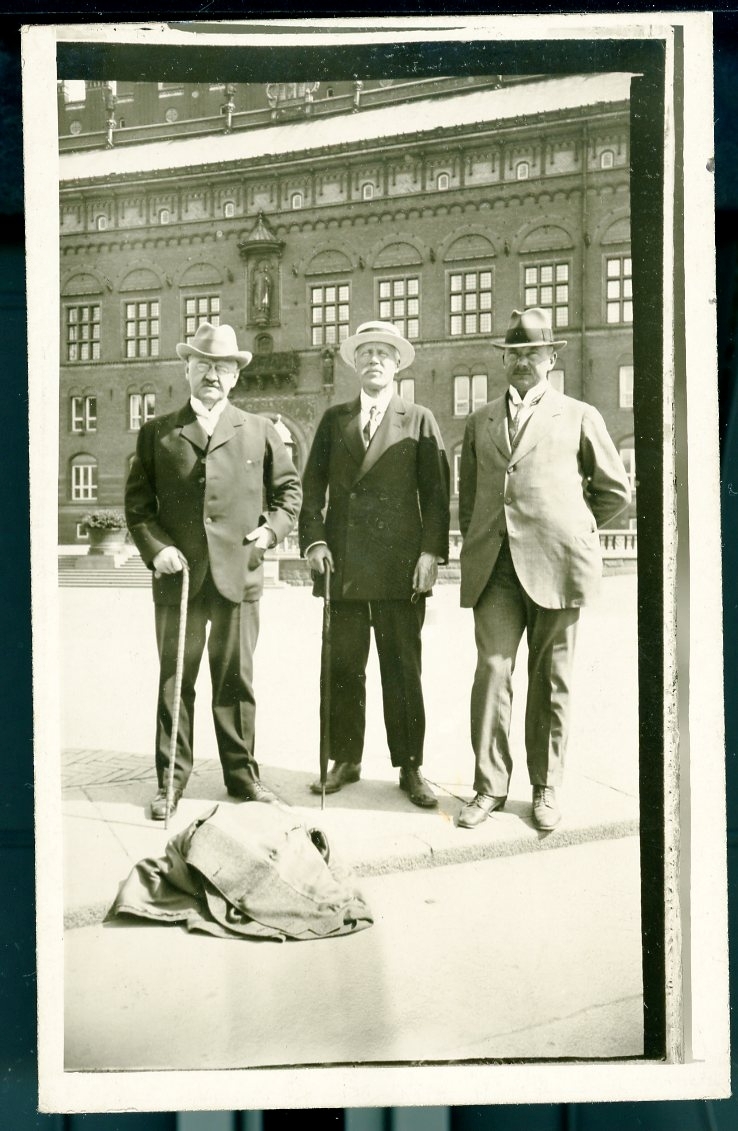 Tre män med hattar och spatserkäppar vända mot fotografen framför rådhuset i Köpenhamn. En el flera rockar ligger slängda på marken framför dem.

Bakpå skrivet: "major E Hedenberg, överste Hjertström, häradshövding C Lundquist. Köpenhamn 1919."
