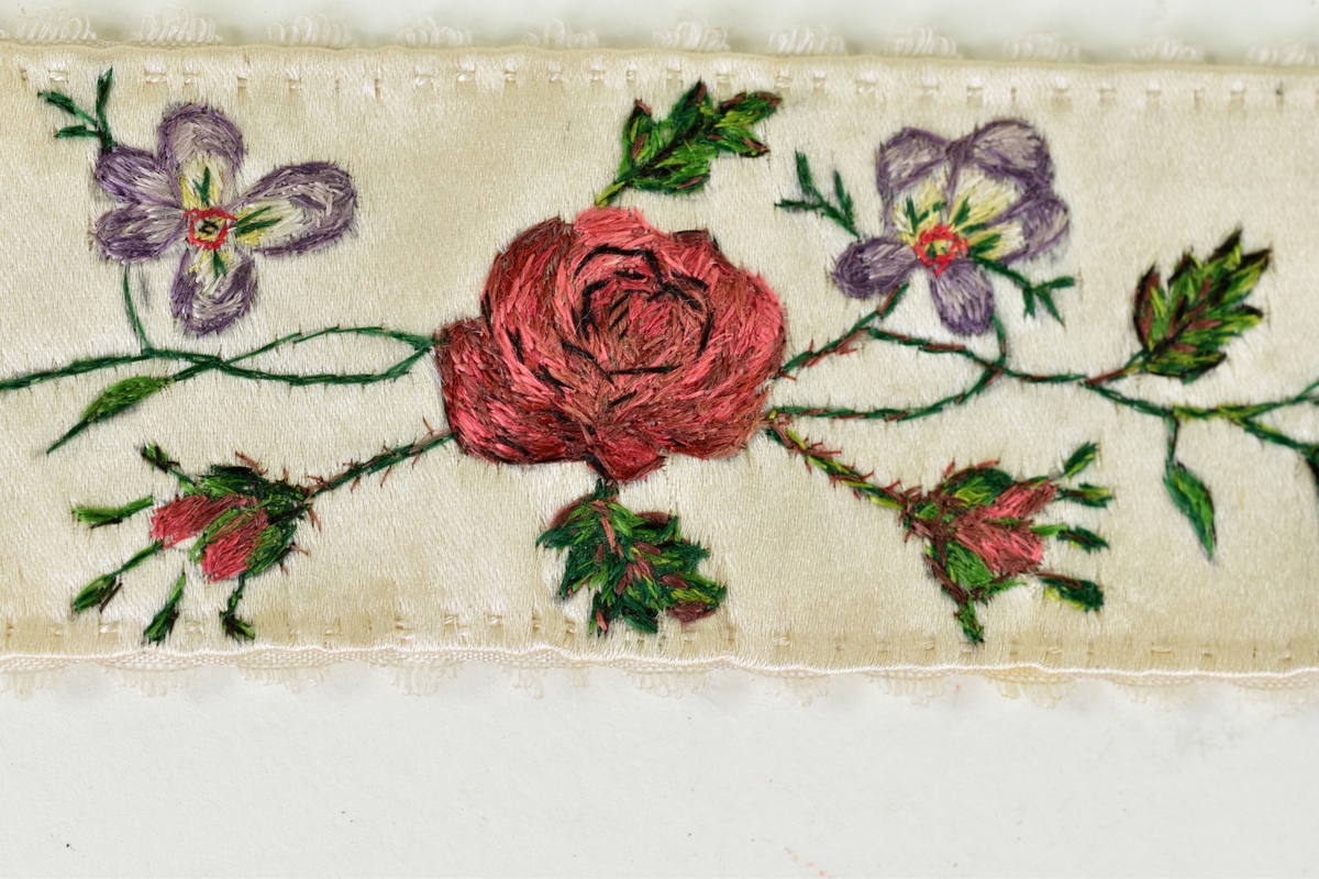Ett par strumpeband av vitt siden med silkebroderier. Broderad blomsterbård med bland annat rosor och violer.