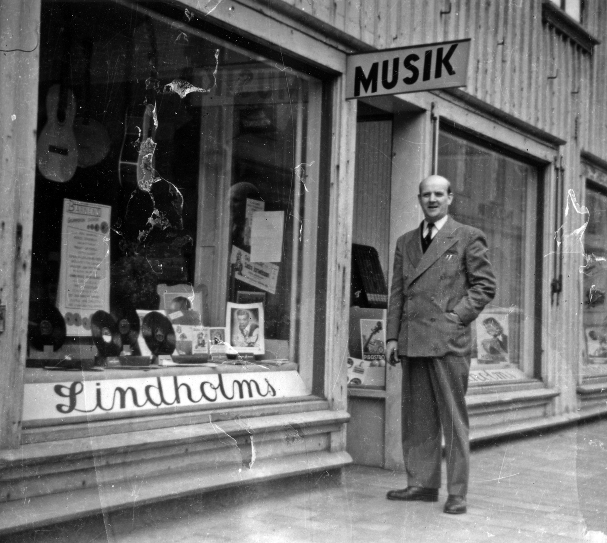 Butiksinnehavare Gunnar Lindholm står utanför sin butik, Lindholms Musikaffär. I skyltfönstret syns både stenkakor och musikinstrument.
Kvarteret Storken 5, Norra Strömgatan.