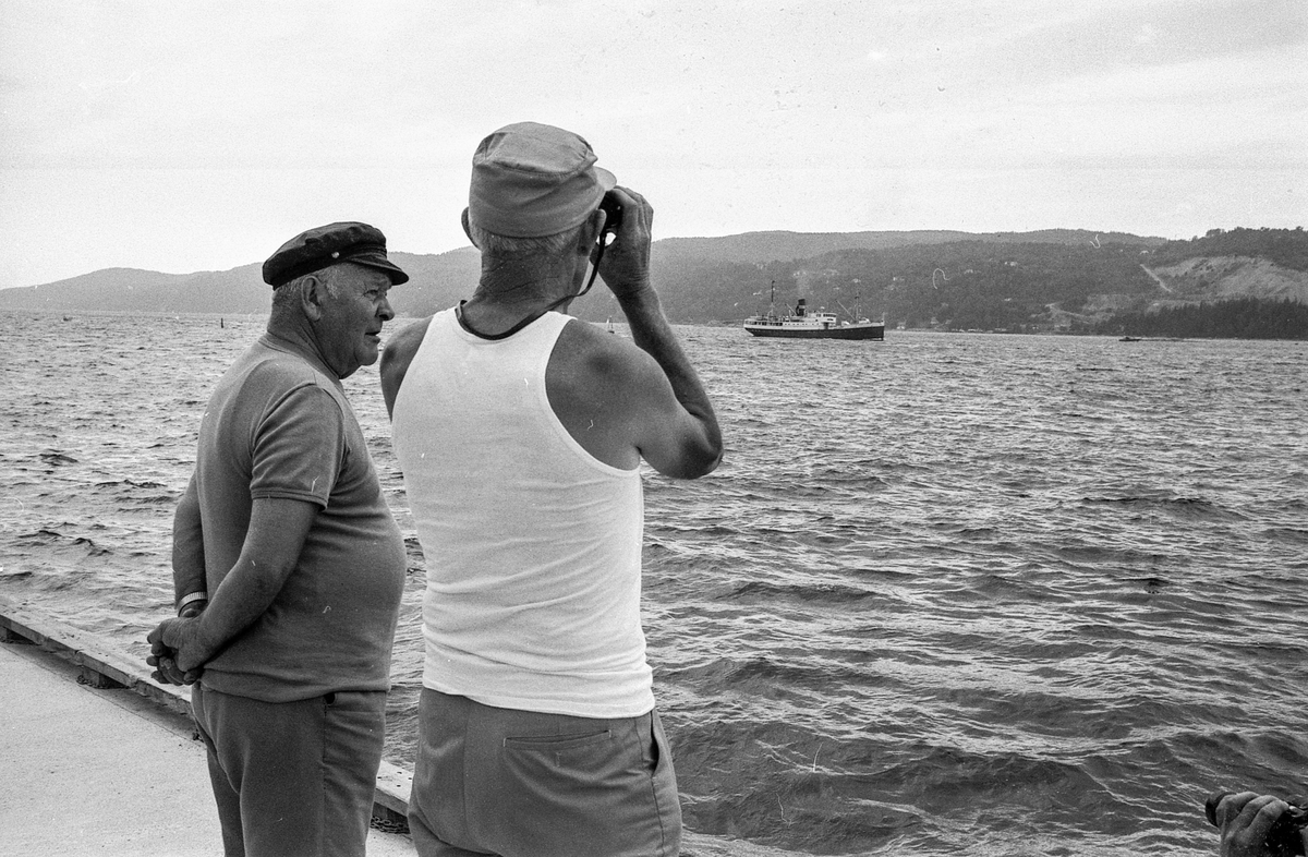 Drøbaksfjorden, sundet ved Oscarsborg. Tre menn på brygga ser på båtene med kikkert. Sandtaket og småhus på Hurum.
Fotograf: ØB Rønning