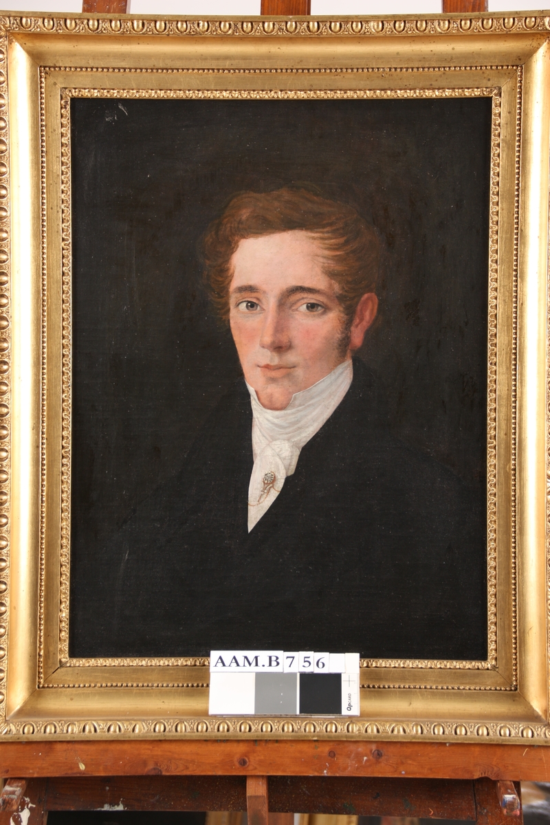 Portrett av ung mann, venstrevendt, foroverbørstet hår, mørk drakt med hvitt halsbind med brosje og lang nål.