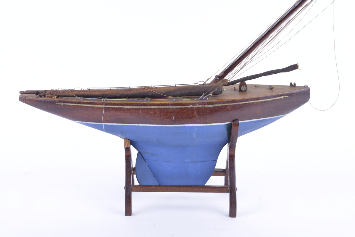 Båtmodell med en hovedmast, samt en mast som er knekt og ligger løst i modellen. Modellen hviler på et stativ av treverk. Ingen seil.