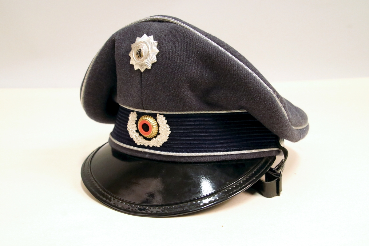 Grå politilue med sort lakkskjerm og detaljer i blått og sølv. Svart hakereim.
Trolig fra 1950/60-tallet.