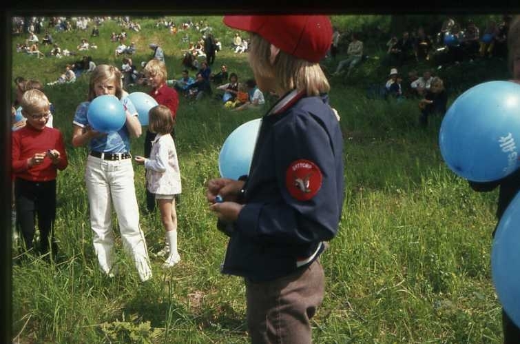 75-årsminnet av Andrées Nordpolsfärd firas med Ballongdagen på Åsavallen i Gränna 1972. Flickor och pojkar som blåser upp blå ballonger. På slänterna sitter publiken. Diabild.