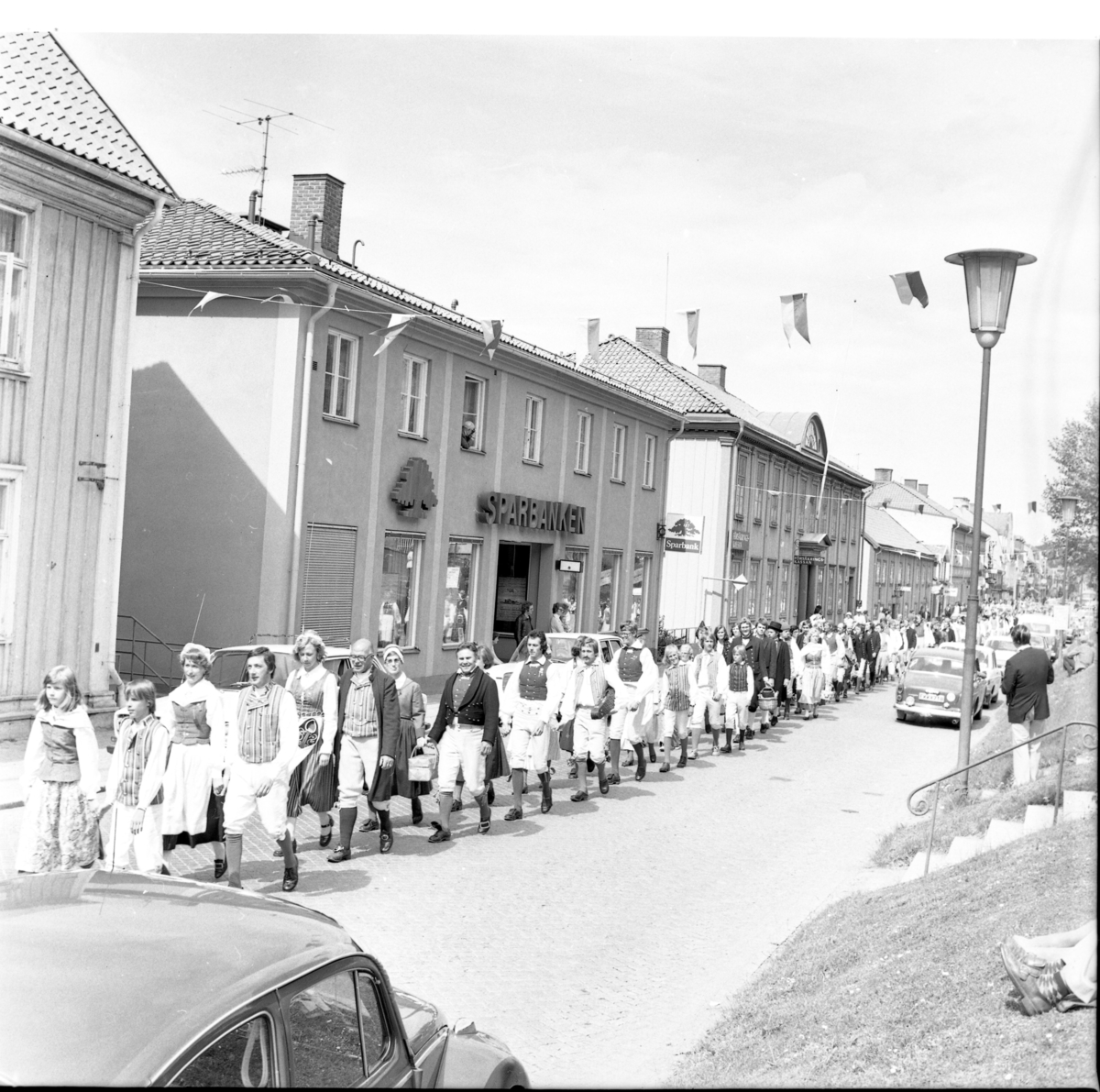 Folkdansare marscherar söderut på Brahegatan i samband med Grännapolkan på Grännaberget, 1975. Till vänster skymtar Sparbanken.