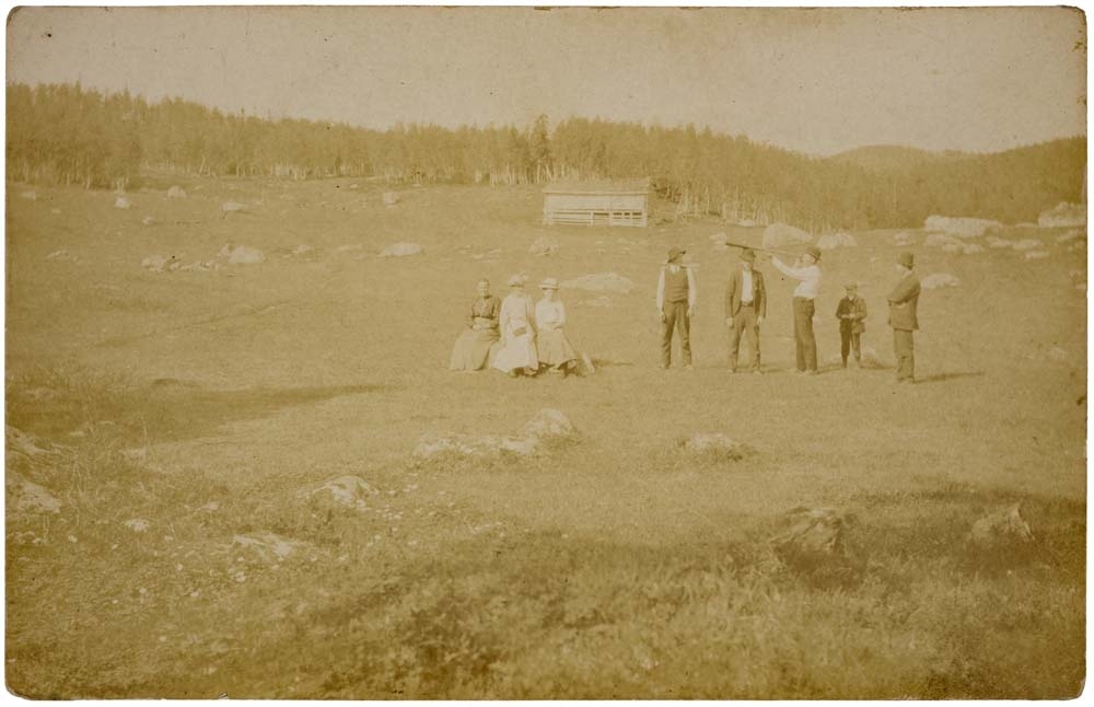 Postkort med gruppebilde av tre kvinner, fire menn og ett barn. Skog, steiner og en bygning i bakgrunnen. Usikkert årstall.