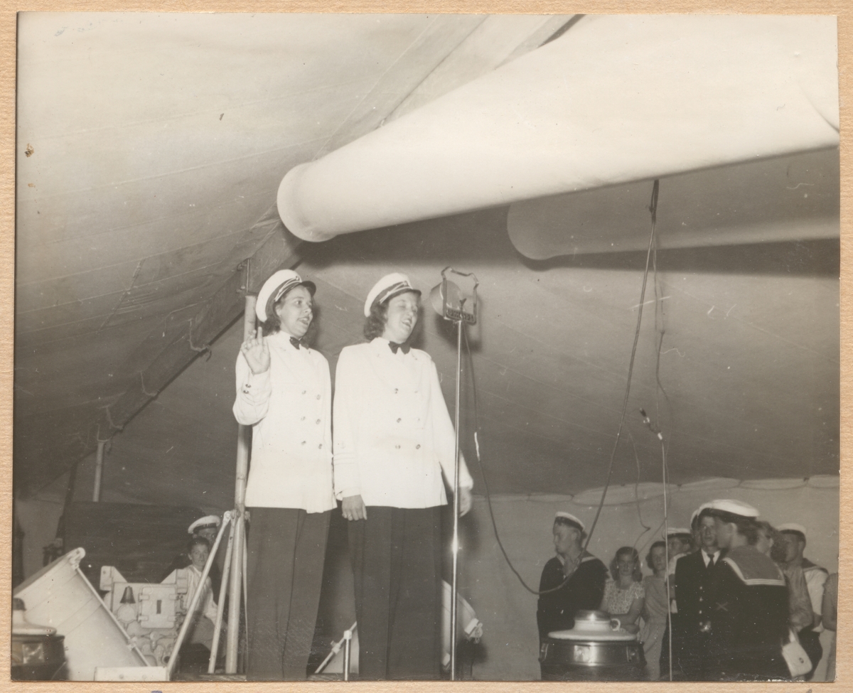 Här underhåller två sjungande damer i uniform de värnpliktiga på Fylgia på en fest. Festen äger rum i ett tält under Fylgias kanoner.