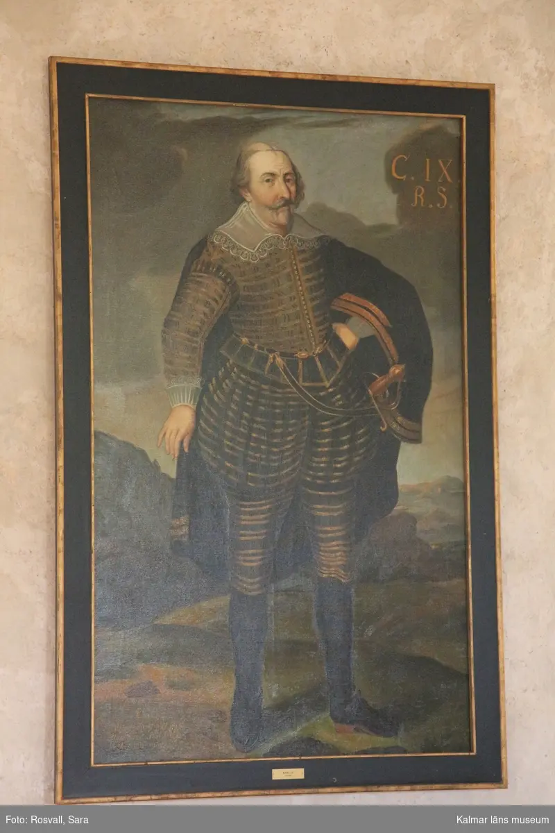 Karl IX i helfigur, stående, i mörk dräkt med guldstickningar, mantel med guldbroderier, spetskrage samt spetsar vid handlederna. Värja, svarta strumpor och skor med spännen.