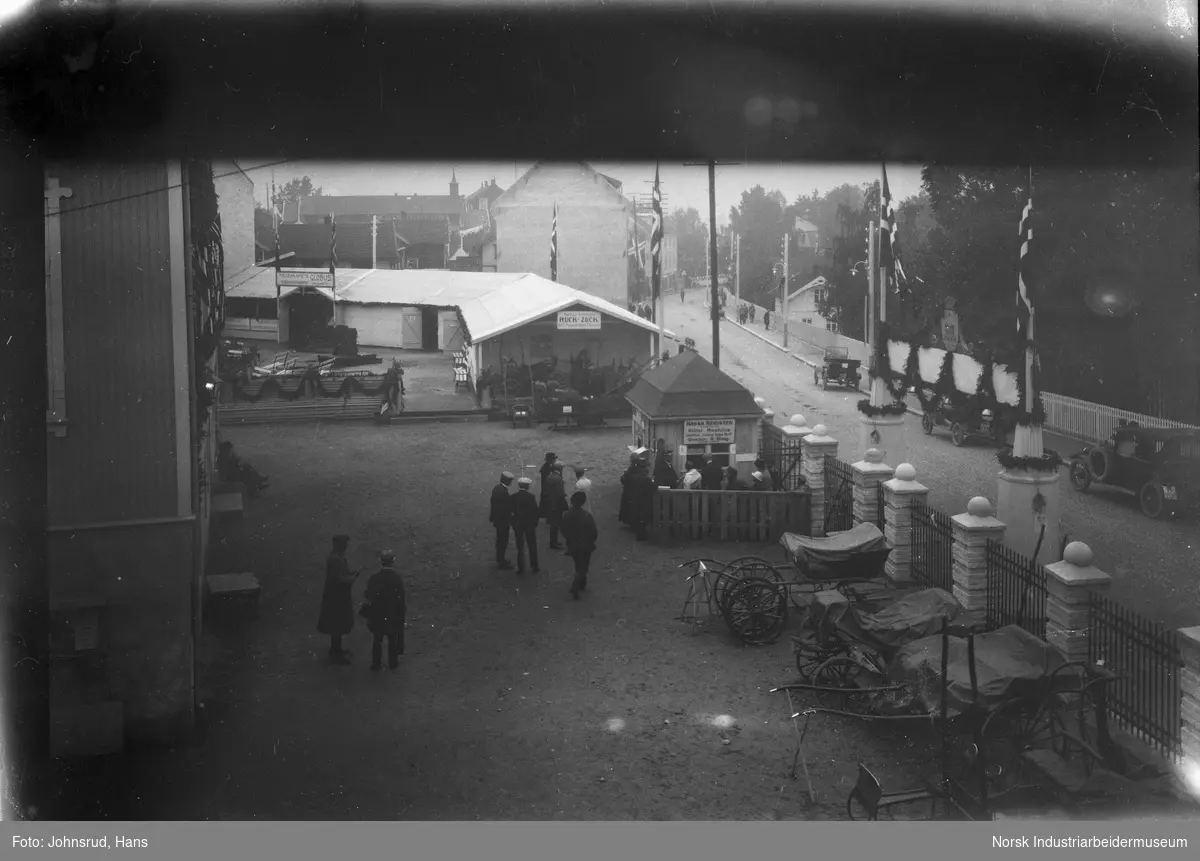 Inngang til Fylkesutstillingen 1922. Biler står parkert i Storgata, mennesker gående i gaten. Hestevogner stående innenfor gjerde ved bygning.