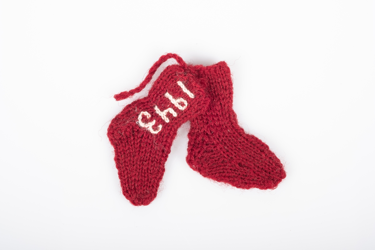 Ett par strikket sokker som er bundet eller sydd sammen. Sokkene er strikket med garn i burgunder. Det er brodert "Grini" på den ene sokken og "1943" på den andre, med hvitt garn.