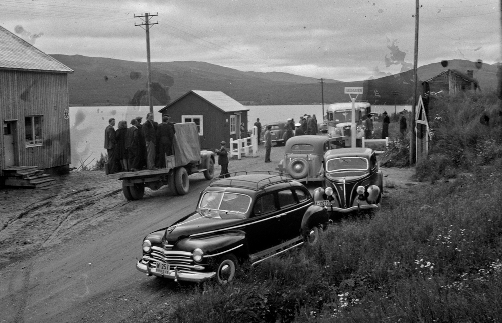 Tollstasjonen ved Joesjø i Sverige, mye folk, biler og busser.
Bilene høyre side, forfra: W-257 Plymouth, sannsynligvis tilhørende Nils Sørum (Drosje),  1946-48 drosje, F-11070 engelskbygd Ford V8 60hk ca. 1937-39, amerikansk Ford V8 1936, førkrigs buss.