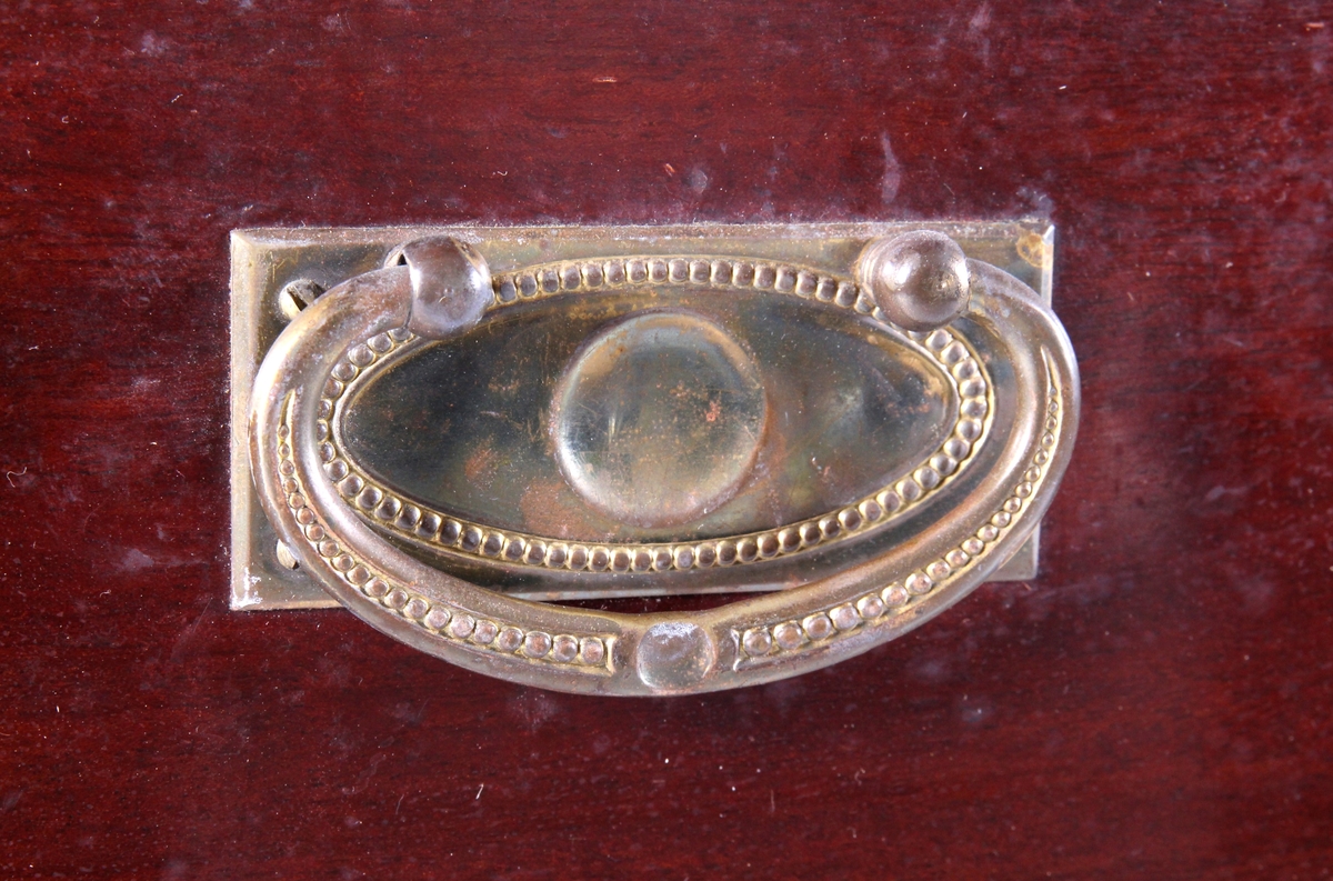 Brunlakkert toalettkommode med ovalformet speil som kan vippes fram og tilbake. Skuffeseksjon med to småskuffer på hver side av speilet. Underdelen består av tre langskuffer som er låsbare, med håndtak og nøkkelskilt