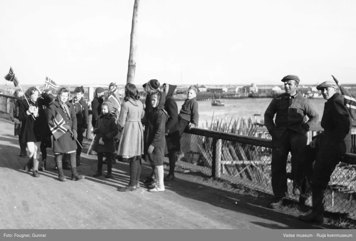 17. mai-tog i Vadsø 1945. Voksne og barn ved et gjerde. Vadsø havn i bakgrunn.