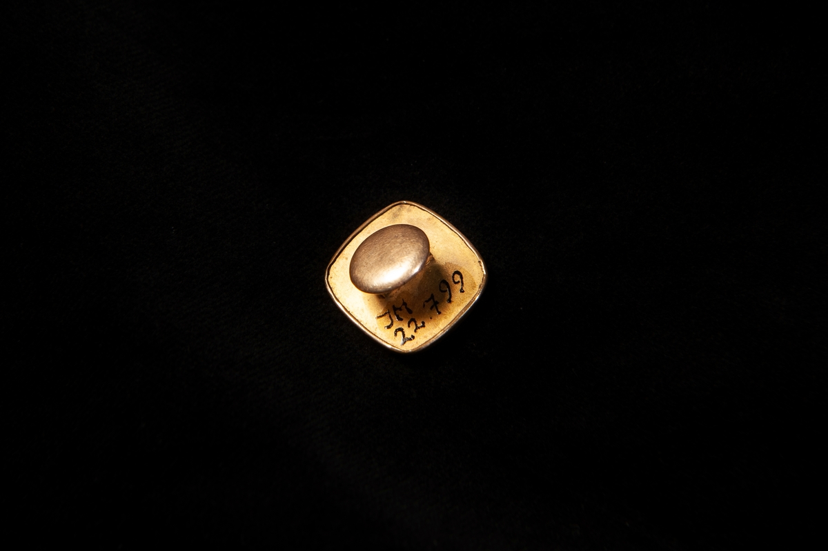 Fyrkantig stolpknapp av förgylld metall (gulddoublé). Präglad dekor i form av zickzack-bårder m.m, och en infattad rund pärlemoknapp 
(troligen imitation) med ett hål i mitten.