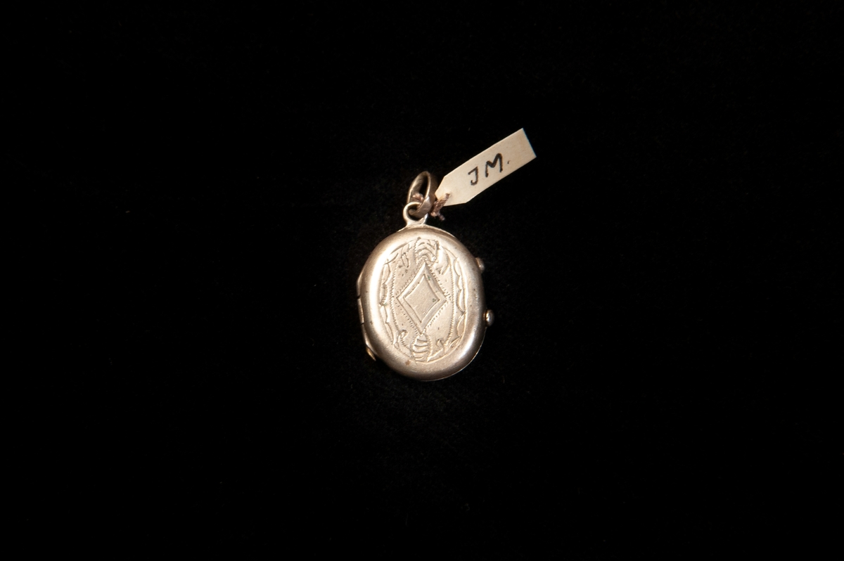 En oval medaljong av tenn(?). Dekorerad med ett graverat hjärta, romb m.m. Medaljongen knäpps med snäpplås. Inuti finns två foton.