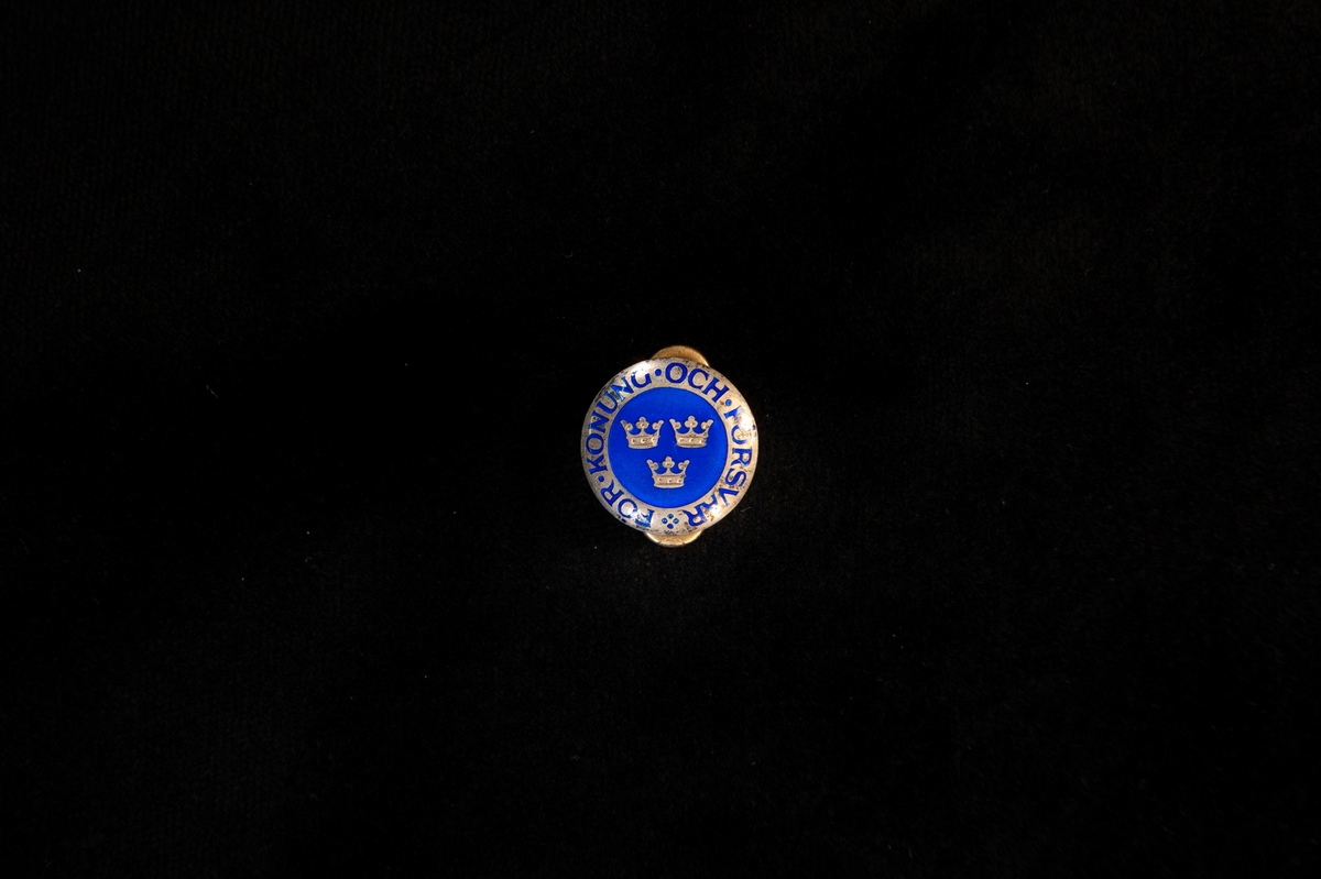 Manschettknapp, rund knapp med emaljdekor i blått och guld, i form av tre kronor med text runt om: "För Konung och Försvar".