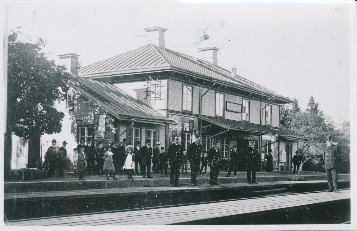 Sommarbild tagen från tidigt 1900-tal på Kilafors Järnvägsstation där flertalet resenäreer och tågpersonal inväntar anklommande tåg. Spåret är ännu inte elektrificerat varpå ånglok brukades.