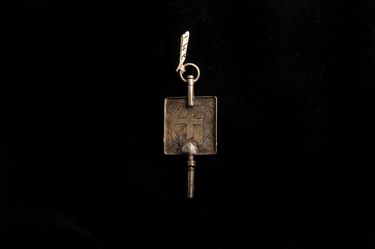 En urnyckel av silver, med fyrkantigt handtag. På ena sidan ett graverat kors och på det andra ett hjärta. Inom hjärtat stämplar.