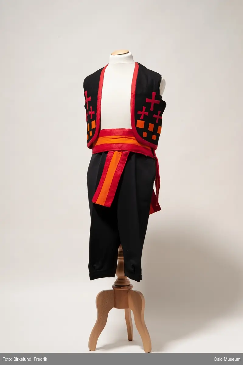 Tredelt svart kostyme.
A: Vest, sort med røde og oransje applikasjoner
B: Belte, bredt oransje/rødt
C: Knebukser