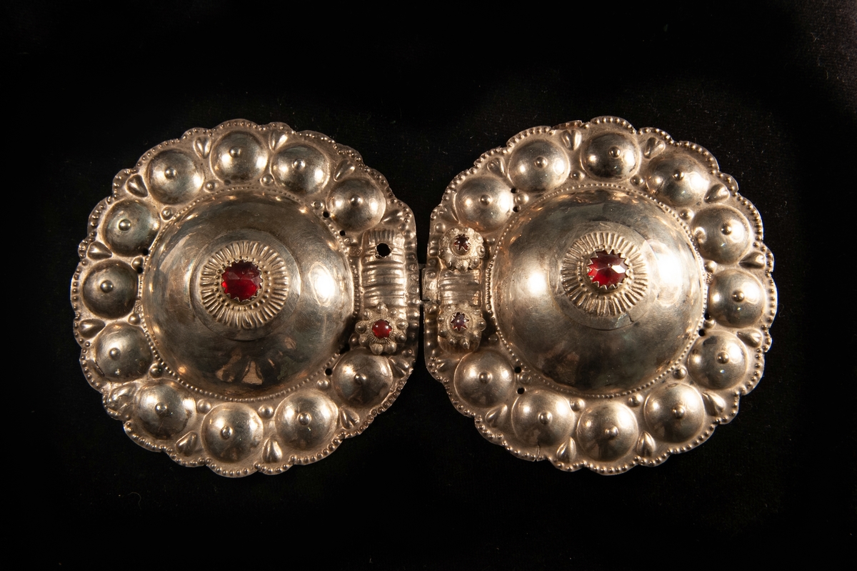 Ett runt tröjspänne av ciselerat silver med 3 infattade stenar av rött glas. De två mindre stenarna infattade gjutna blommor i filigranstil. Har en hake som fästes i en hyska i JM.19963:2.