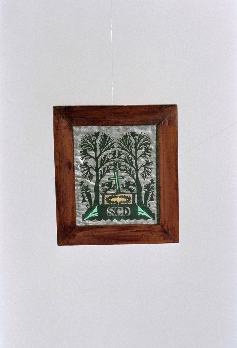 Minnestavla i glas och slät, brun ram, silhuettklippt motiv i form av två träd, fåglar, ett kors, initialern"S C D". Svart papper mot silverpapper.