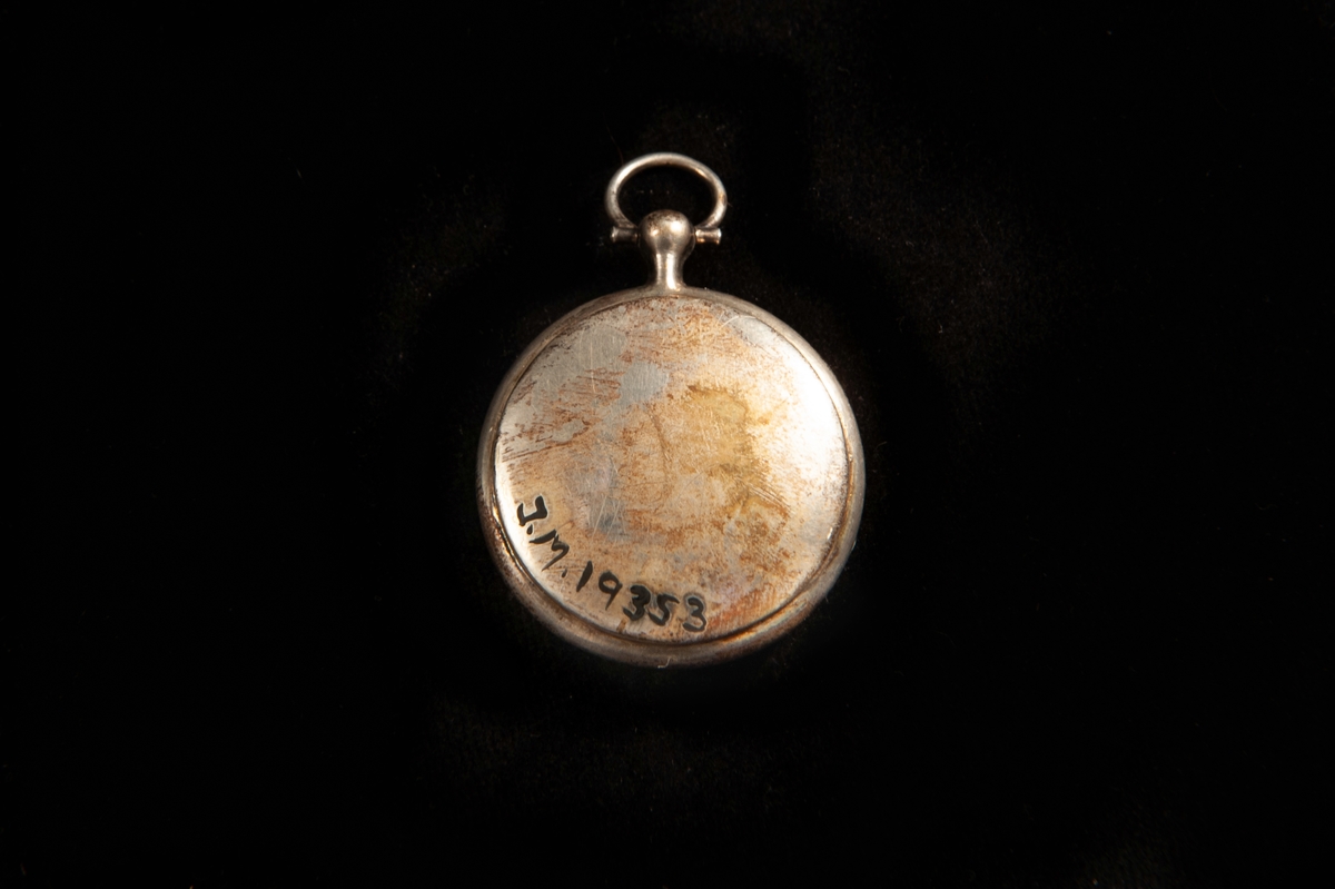 Medaljong i form av en klockboett. Stämplar invändigt på locket: . Släta sidor, en knopp upptill med en oval ring att hänga kedjan i. Invändigt förgylld.