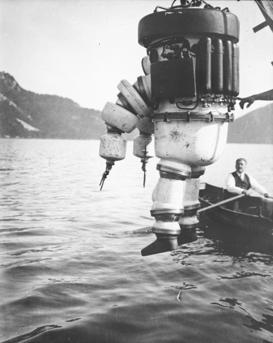 Dykker i panserdrakt heises ned i vannet, mann i robåt til høyre i motivet.