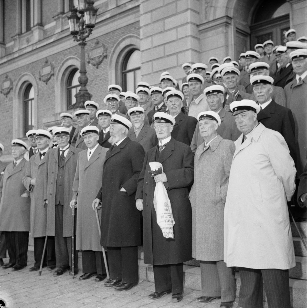 Flera personer iförda studentmössor står på trappan vid Universitetetshuset, Östgöta nations minneslandskap, Uppsala 1949