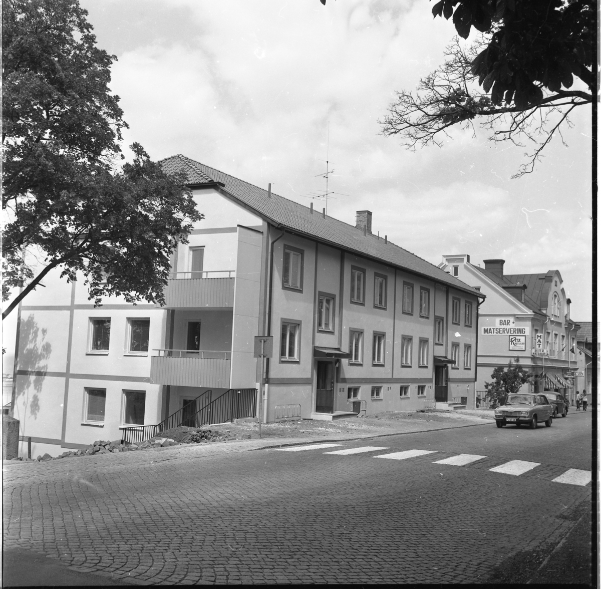 Nybyggt hyreshus på Brahegatan 1 i Gränna. Längre ner på gatan skyltas för Ritz Matsalar, mat och polkagrisar. En bil (Simca?) är på väg ut ur staden.