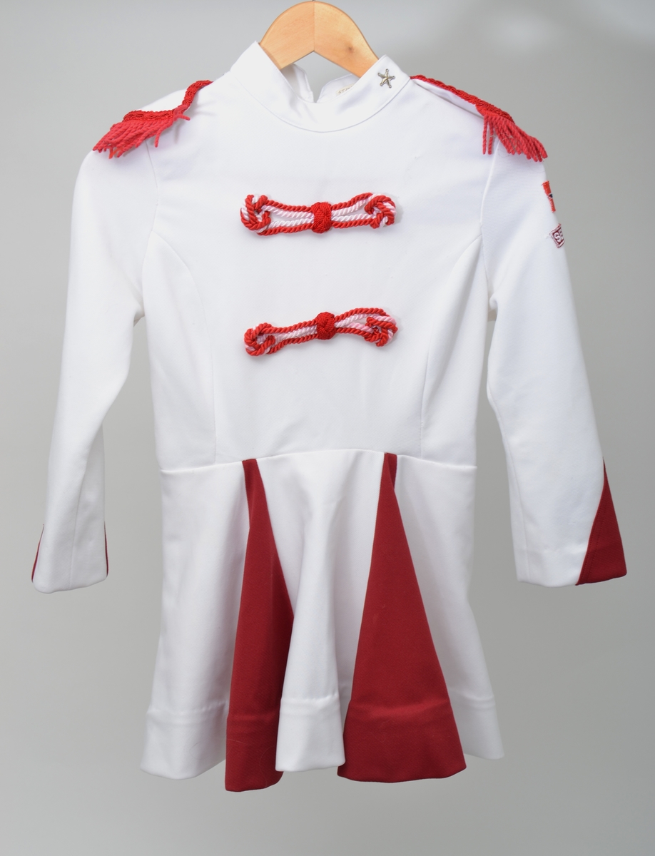 Kort hvit og rød uniformskjole med glidelås i ryggen. Skjøret er sydd sammen av vekselsvis røde og hvite kileformede tøystykker. Dekorert med snorer og epåletter.