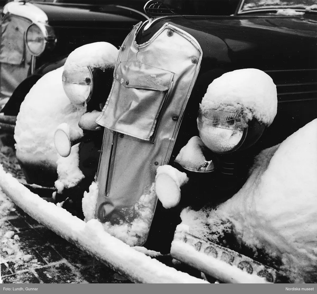 Snö ligger på fronten av en Plymouth De Luxe av 1936 års modell. Kylaren har en avskärmning påsatt för att hålla motorn varm.