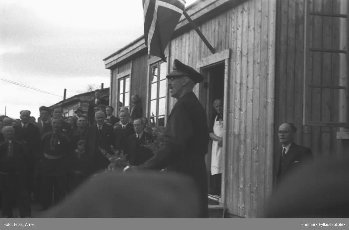 Kongebesøk i Langnes juli 1946. Kong Haakon VII reiste landet rundt for å se på krigsødeleggelsene og gjenreisningsarbeidet. Kongen holder en tale utenfor Tana Turiststasjon. Bland publikum, venstre side fra Kongen fylkesmann Hans Gabrielsen, og høyre side Per Fokstad, ordfører i Tana