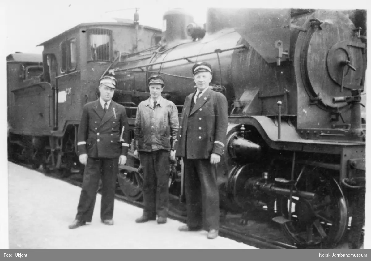Lokomotivfører Eberhard Hasselaas (til høyre) og fyrbøter Rolf Søia (i midten) foran damplokomotiv type 21a nr. 207 på Kongsberg stasjon.