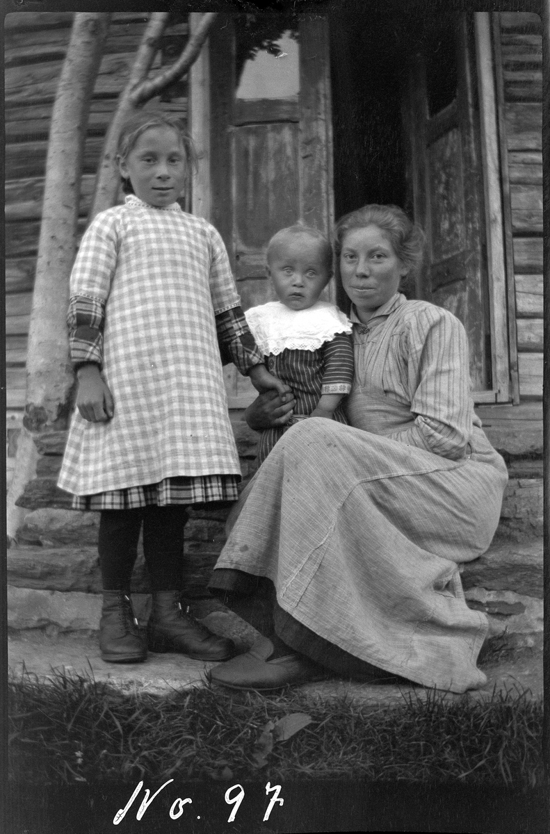 Portrett av en kvinne og to barn sittende på steintrappa til en laftet bygning.