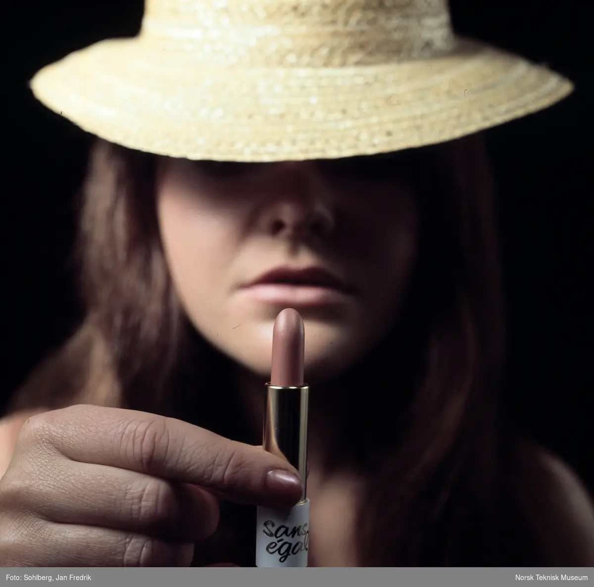 Reklamefoto for leppestift. Nærbilde av kvinne med hatt som holder en leppestift opp foran ansiktet sitt.