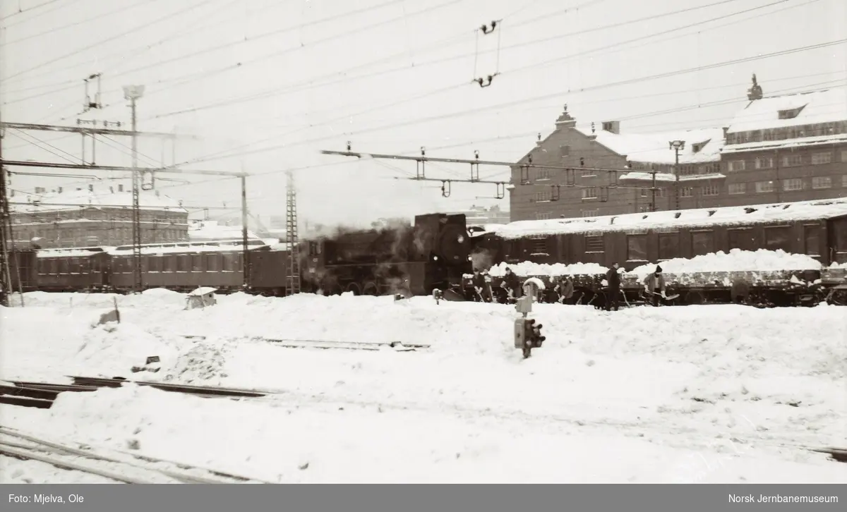 Damplokomotiv type 31b nr. 451 med godsvogner litra N2 lastet med snø på Oslo Østbanestasjon. Mannskaper spar sne opp i vognene med håndmakt.