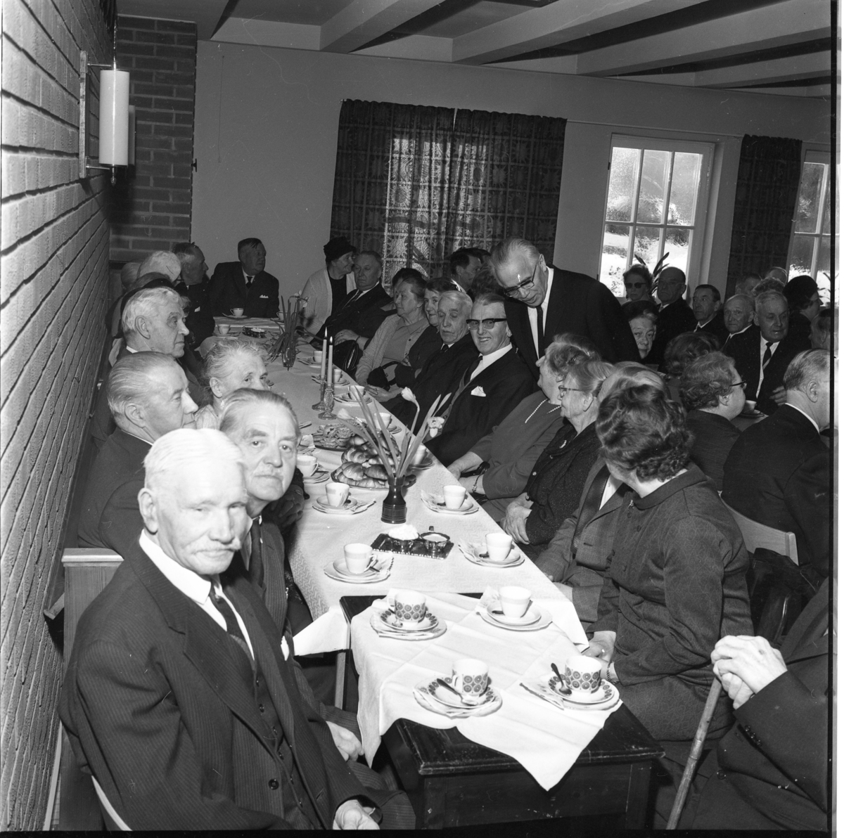 En samling äldre män och kvinnor sitter vid dukade kaffebord, troligen i Filadelfiakyrkan i Gränna. Närmast till vänster sitter "Kalle på Rhen". Mannen som står upp hukad över bordet är Robert Svensson, pastor i församlingen i Gränna.
