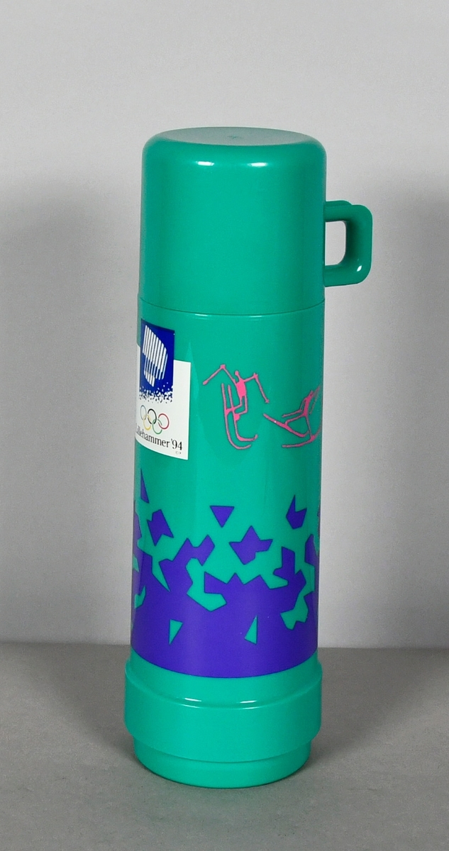 Grønn termos med kopp. Temosen har snøkrystallmønster i blått og piktogrammer i rosa, og nordlyslogo for Lillehammer '94 og de olympiske ringene i farger. Termosen rommer 0,5 l.